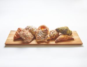 A wooden board displays an assortment of pastries, including croissants and danishes, each topped with various glazes, nuts, and powdered sugar.