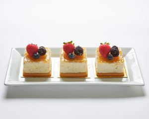 Three square layered desserts topped with strawberries, blackberries, blueberries, and a glossy glaze are arranged in a row on a rectangular white plate.
