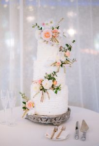 Three-tiered white wedding cake decorated with pastel flowers on a silver stand, with two champagne flutes, a cake server, and napkin with forks beside it.