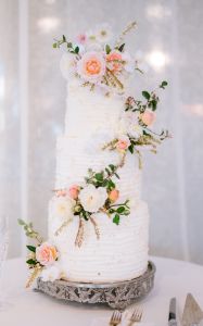 Three-tiered white wedding cake decorated with pink and white flowers, displayed on a silver stand with champagne flutes and cutlery on the table nearby.
