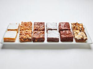 An assortment of desserts beautifully arranged on a platter, featuring lemon bars, nut bars, powdered sugar-topped chocolate brownies, plain chocolate brownies, and marshmallow-topped brownies.