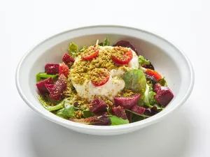 A white bowl filled with Burrata cheese with mixed greens, beets, tomatoes, and crushed pistachios, dressed in extra-virgin olive oil, vinegar and truffle sea salt.
