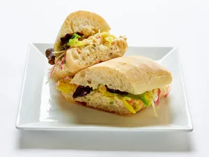 A sandwich of smashed chickpeas with greens, carrots, radish, sweet cherry peppers, and vegan mayonnaise on ciabatta bread, cut in half and served on a white square plate.