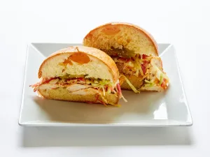 A Buffalo chicken sandwich with avocado, coleslaw, and ranch dressing, cut in half on a square white plate.