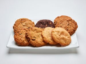 The Classic Cookie Platter: Assorted features a white rectangular plate with seven cookies, including chocolate chip, oatmeal, and dark chocolate cookies, arranged in a circular pattern.