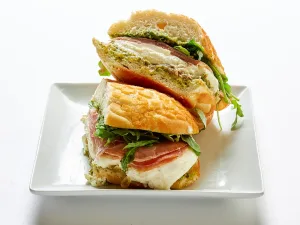 A Prosciutto and burrata cheese sandwich with wild arugula, Balsamic onions, and pesto on ciabatta bread, cut in half and stacked on a white square plate.