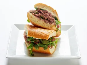 Two halves of the Roast beef, wild arugula & truffle-ricotta cheese spread sandwich on a baguette are stacked on a white square plate.
