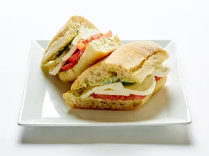 Grilled fresh mozzarella and tomatoes, fresh basil, and pesto aioli on ciabatta bread, served halved on a square white plate.