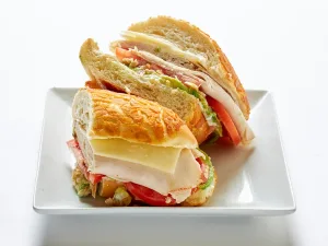 A Grilled Turkey Sandwich with avocado, white cheddar cheese, balsamic onions, tomatoes, and mayonnaise on a crusty roll, cut in half and served on a white plate.