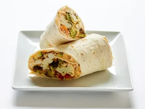 A Roasted Veggie Wrap, cut in half on a white square plate, is filled with broccoli, zucchini, bell peppers, cauliflower, onions, and roasted red pepper hummus with zahtar, all rolled in vegan lavash flat bread.