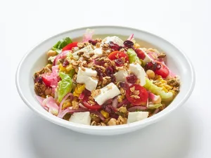 A white bowl filled with chopped salad featuring garbanzo beans, roasted corn, feta cheese, red onions, tomatoes, savory granola, dried cranberries, and mustard-dill vinaigrette on a white background.
