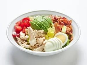 A white bowl holds a Cobb salad with chicken, bacon, avocado, blue cheese, tomatoes, eggs, and Ranch dressing in San Francisco on a white background.