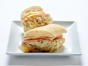A ciabatta sandwich with grilled albacore tuna, capers, white cheddar cheese, tomatoes, and balsamic onions, cut in half and served on a white square plate.