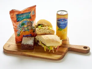 The Smashed Chickpea Sandwich Box includes a sandwich, brownie, Hawaiian BBQ chips, and Sanpellegrino Limonata on a wooden board.