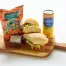 The Smashed Chickpea Sandwich Box includes a sandwich, brownie, Hawaiian BBQ chips, and Sanpellegrino Limonata on a wooden board.