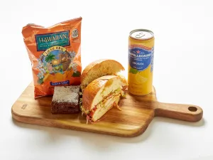 The Buffalo Chicken Sandwich Box includes a sandwich, brownie, Hawaiian BBQ chips, and a can of San Pellegrino Limonata, all presented on a wooden serving board.