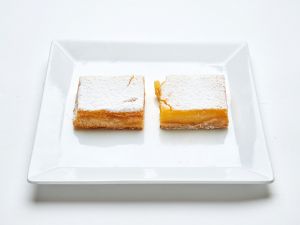 Two Lemon Bars with powdered sugar are displayed on a square white plate set against a white background.