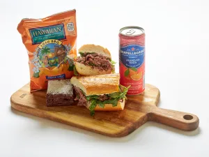 The Roast Beef Sandwich Box includes a roast beef sandwich with greens, a brownie, Hawaiian BBQ chips, and a can of Sanpellegrino soda, all presented on a wooden serving board.