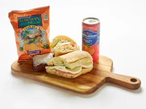 The Mozzarella & Tomatoes Sandwich Box includes a sandwich, mini sandwich, brownie, Hawaiian BBQ chips, and a can of San Pellegrino Aranciata Rossa, all served on a wooden board.