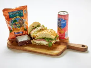 The Prosciutto & Burrata Cheese Sandwich Box includes a halved sandwich, a brownie, Hawaiian BBQ chips, and a can of San Pellegrino sparkling drink.