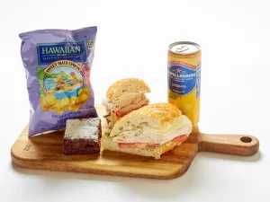 The Turkey & Avocado Sandwich Box includes a halved sandwich, brownie, Maui onion chips, and a San Pellegrino lemon soda, all served on a wooden board against a white background.