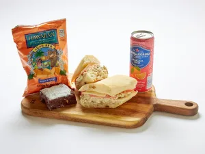 A wooden board featuring an Albacore Tuna Sandwich Box, San Pellegrino can, Hawaiian BBQ chips, and a brownie.