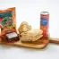 A wooden board featuring an Albacore Tuna Sandwich Box, San Pellegrino can, Hawaiian BBQ chips, and a brownie.