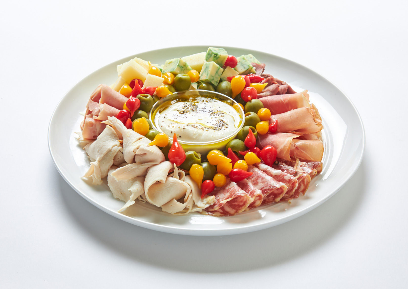 A white plate with assorted cured meats, cheeses, and colorful peppers arranged around a bowl of creamy dip in the center—perfect for sharing at parties or adding to your favorite platters.