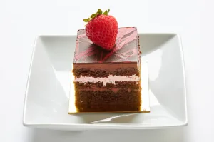 A square piece of Chocolate Strawberry cake with rich chocolate frosting and a whole strawberry, served on a white plate—perfect for pairing with creamy cheesecake.