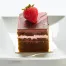 A square piece of Chocolate Strawberry cake with rich chocolate frosting and a whole strawberry, served on a white plate—perfect for pairing with creamy cheesecake.