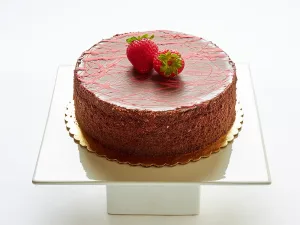 Chocolate cake with a smooth, glossy top, decorated with red drizzle and two whole strawberries, displayed on a white cake stand—a delightful addition to our Cakes Menu.