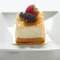 A square piece of Coconut Pineapple cheesecake, topped with a strawberry, blueberry, and blackberry, served on a white plate.