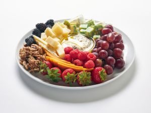 A white plate, styled like elegant platters, features strawberries, raspberries, red grapes, blackberries, cheese cubes, cucumber, brie, walnuts, and a dish of honey arranged in neat sections.