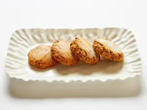 Four round gluten-free cookies are arranged in a row on a rectangular, wavy-edged white plate against a white background, creating an inviting Gluten-Free Cookie Platter: Assorted display.