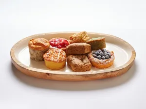 Our Gluten-Free Morning Pastry Platter offers eight assorted pastries and cakes, such as muffins, fruit tarts, and brownies, beautifully displayed on an oval wooden tray atop a white surface.