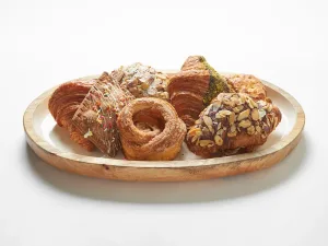 A wooden platter holds an assortment of pastries, including croissants, danishes, and rolls, topped with nuts, icing, and sprinkles on a white background.