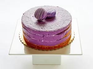 A round, single-tier cake with purple frosting and two purple chocolate truffles on top, displayed on a white square cake stand—a delightful choice from our Cakes Menu.