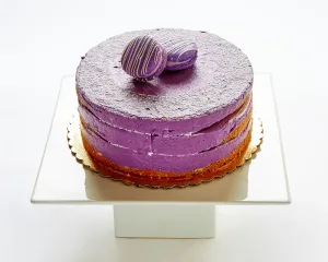 A round, single-tier cake with purple frosting and two purple chocolate truffles on top, displayed on a white square cake stand—a delightful choice from our Cakes Menu.