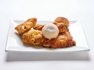 The Vegan Morning Pastry Platter is displayed on a white rectangular plate, featuring a powdered sugar-dusted round pastry, turnovers, and assorted vegan baked goods against a white background.