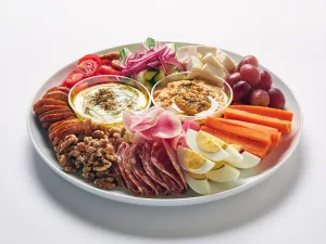 The Mezze Platter features a white plate with sliced vegetables, grapes, hard-boiled eggs, walnuts, cured meats, crackers, pickled onions, roasted vegetable pasta with pesto and Parmesan cheese, plus two dips.
