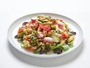 The Pasta Salad Platter features penne, cherry tomatoes, green olives, mixed greens, pesto, and grated Parmesan cheese served on a white plate.