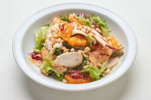 A white plate of Mixed Greens with tomatoes & mustard-dill vinaigrette, topped with sliced chicken breast, orange segments, and pine nuts on a white background.
