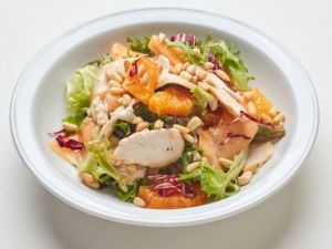 A white plate of Mixed Greens with tomatoes & mustard-dill vinaigrette, topped with sliced chicken breast, orange segments, and pine nuts on a white background.